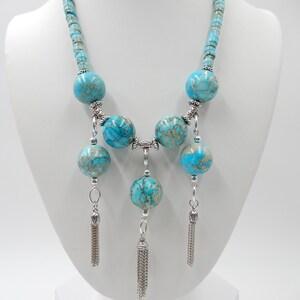 Pu&ograve; includere: Una collana di dichiarazione blu turchese con grandi perline rotonde e accenti argentati. La collana ha tre nappe argentate che pendono dal fondo.