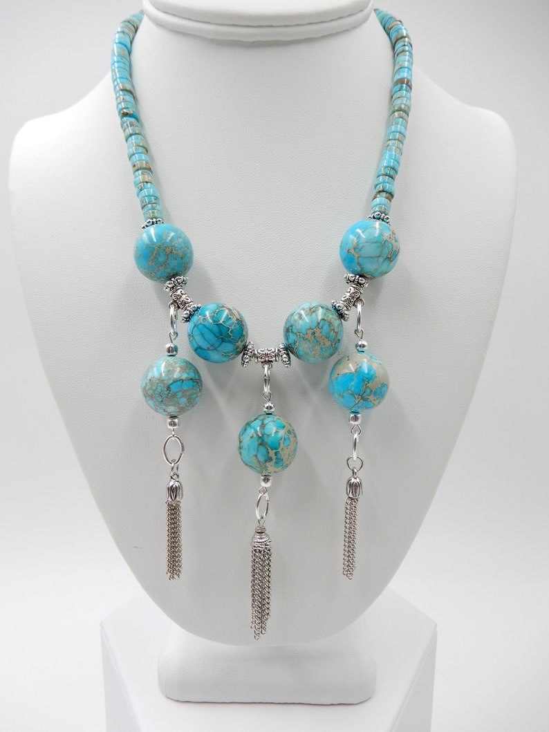 Pu&ograve; includere: Una collana di dichiarazione blu turchese con grandi perline rotonde e accenti argentati. La collana ha tre nappe argentate che pendono dal fondo.