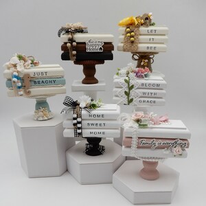 May include: Six white wooden tiered stands with stacked books decorated with various embellishments, including flowers, ribbons, and beads. The books have different sayings, including "Just Beachy", "This is my happy Home", "Let it Bee", "Bloom with Grace", "Home Sweet Home", and "Family is everything".