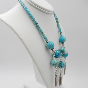 Pu&ograve; includere: Una collana blu turchese con grandi perline rotonde e nappe d'argento. La collana &egrave; su un manichino bianco.