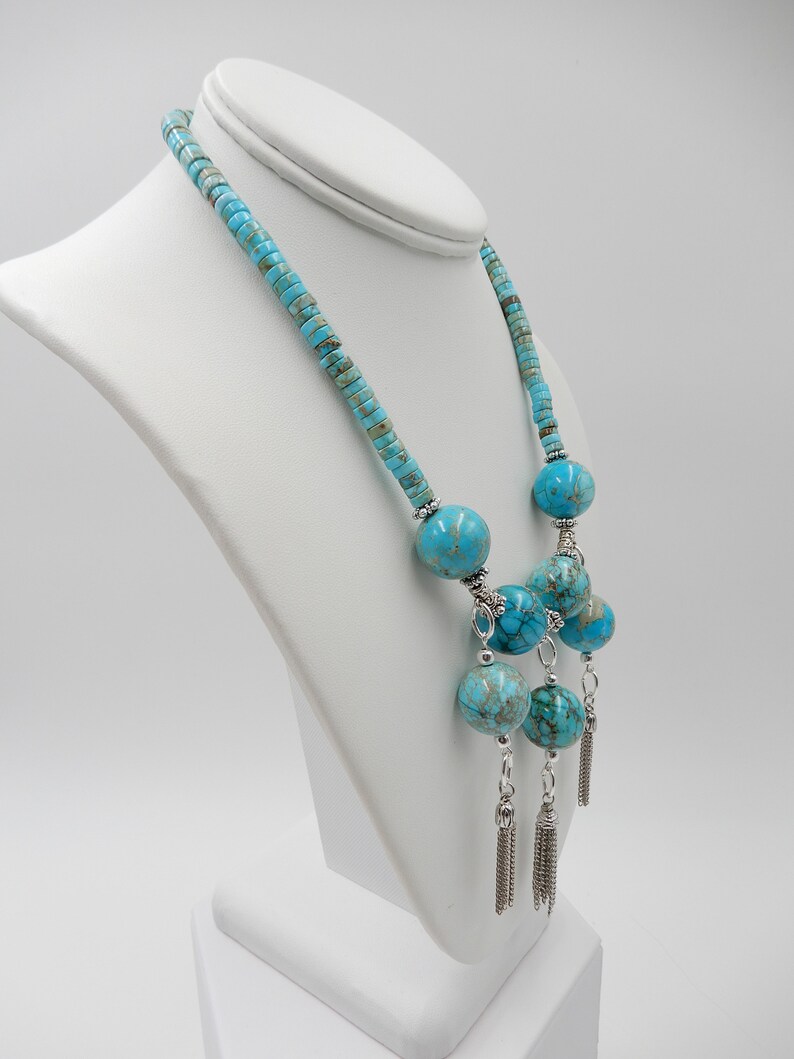 Pu&ograve; includere: Una collana blu turchese con grandi perline rotonde e nappe d'argento. La collana &egrave; su un manichino bianco.
