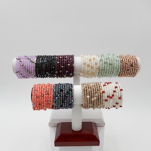 May include: A display of colorful beaded bracelets in various colors, including purple, black, maroon, cream, green, copper, coral, and white. The bracelets are stacked on a white stand with a red base.