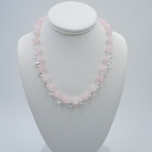 May include: A pink and clear crystal necklace with round beads and teardrop accents. The necklace is displayed on a white mannequin.