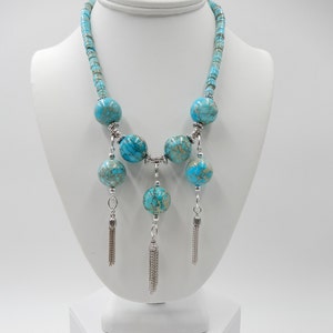 Pu&ograve; includere: Una collana turchese e argento con una lunga catena e tre nappine. La collana presenta grandi perle rotonde turchesi con accenti argentati.