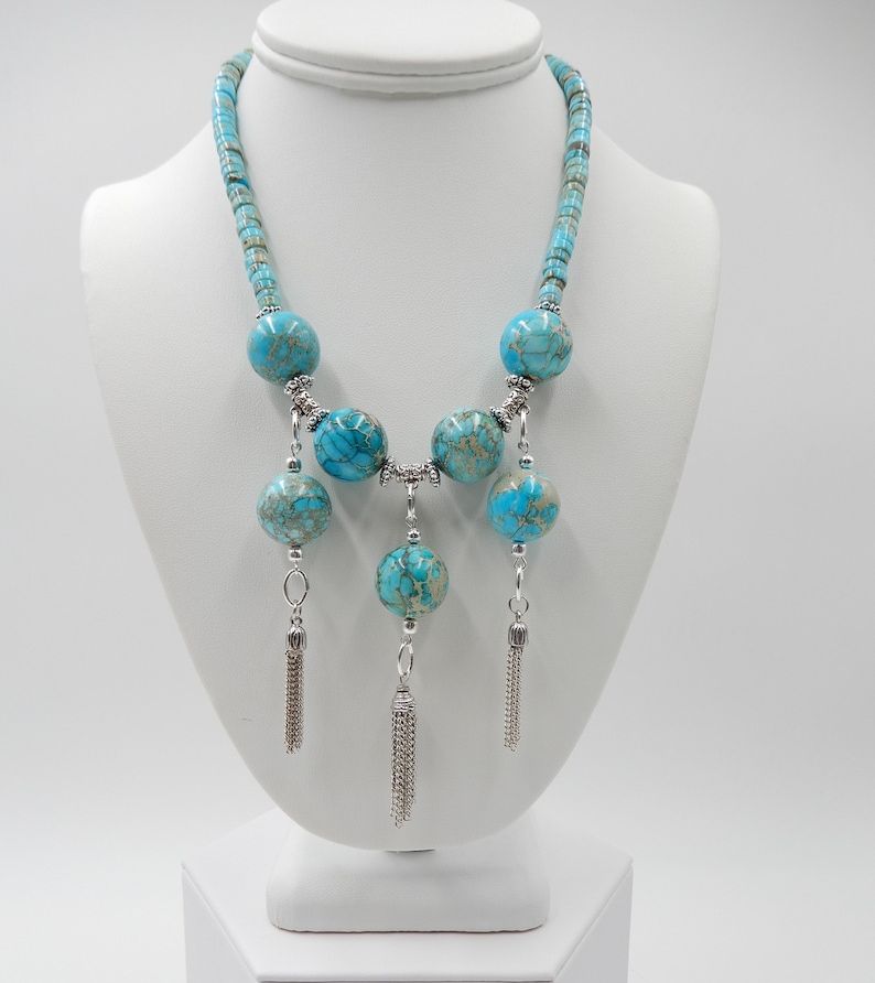 Pu&ograve; includere: Una collana turchese e argento con una lunga catena e tre nappine. La collana presenta grandi perle rotonde turchesi con accenti argentati.