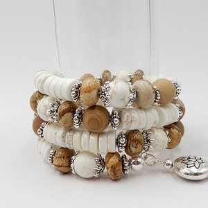 May include: A multi-strand bracelet with white and brown beads and silver accents. The bracelet has a silver charm with a floral design.
