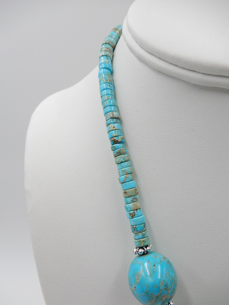 Pu&ograve; includere: Una collana turchese e argento con un grande ciondolo rotondo di turchese in basso. La collana &egrave; composta da piccole perle quadrate di turchese.