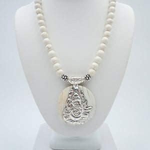 May include: White howlite bead necklace with a silver pendant featuring an intricate design. The pendant is oval shaped and has a white howlite inlay.