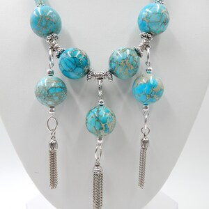 Pu&ograve; includere: Una collana turchese e argento con un pendente lungo a forma di nappa. La collana &egrave; composta da perle rotonde turchesi con accenti argentati e tre lunghe nappe argentate.