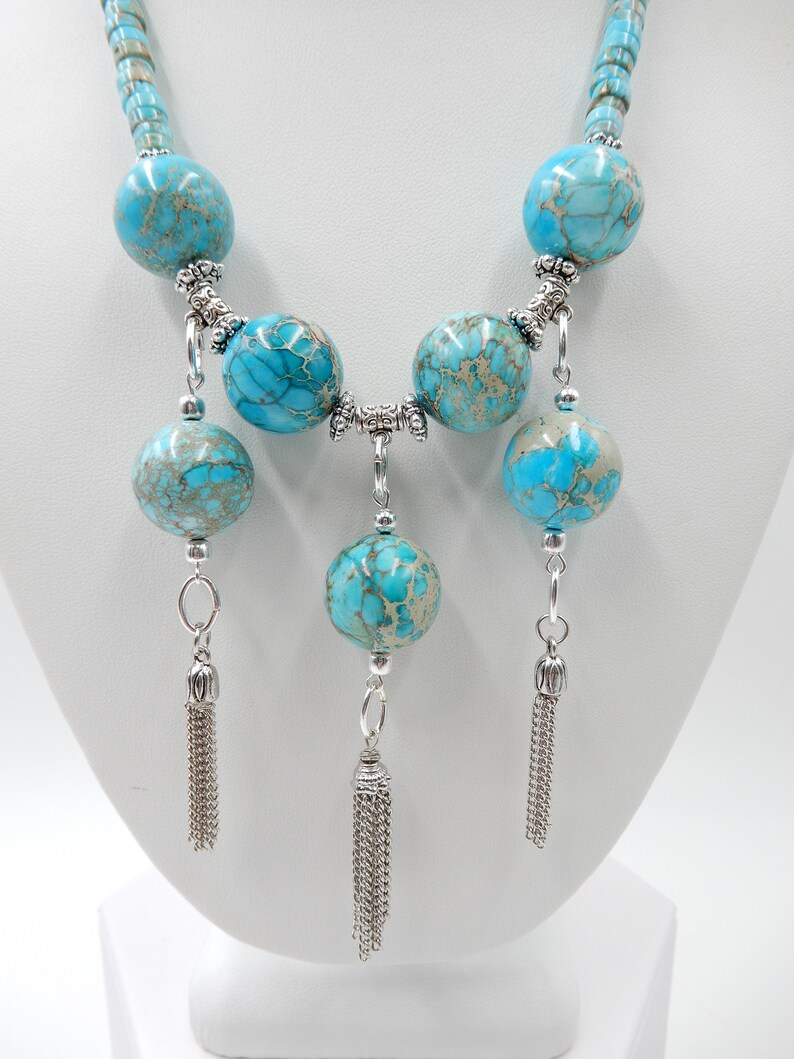 Pu&ograve; includere: Una collana turchese e argento con un pendente lungo a forma di nappa. La collana &egrave; composta da perle rotonde turchesi con accenti argentati e tre lunghe nappe argentate.