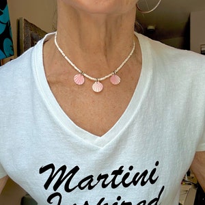 Puede incluir: Una camiseta blanca con una inscripci&oacute;n en cursiva negra que dice "Martini Inspired". La persona lleva un collar de cuentas blancas con tres colgantes de conchas rosas.