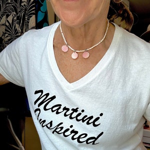 Puede incluir: Camiseta blanca con el texto "Martini Inspired" en escritura cursiva negra. La persona lleva un collar de cuentas blancas con tres dijes de conchas rosadas.