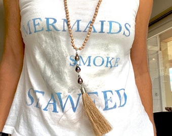 Puede incluir: Una camiseta blanca de tirantes con el texto "MERMAIDS SMOKE SEAWEED" en letras azules. La persona lleva un collar largo de cuentas marrones y blancas con un borla y perlas oscuras.