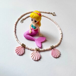 Puede incluir: Un collar de cuentas blancas con tres colgantes de conchas rosadas. El collar est&aacute; decorado con peque&ntilde;as cuentas doradas y diamantes de imitaci&oacute;n.
