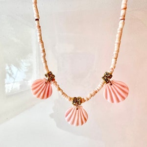 Puede incluir: Un collar de cuentas blancas con tres colgantes de concha rosa. Cada colgante de concha tiene un acento dorado y diamantes de imitaci&oacute;n transparentes.