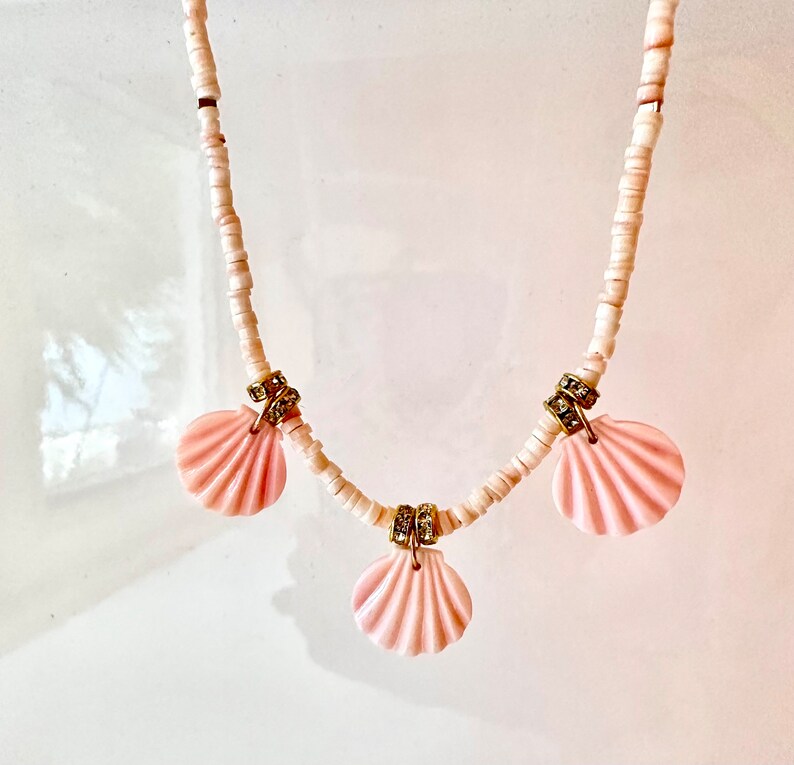 Puede incluir: Un collar de cuentas blancas con tres colgantes de concha rosa. Cada colgante de concha tiene un acento dorado y diamantes de imitaci&oacute;n transparentes.