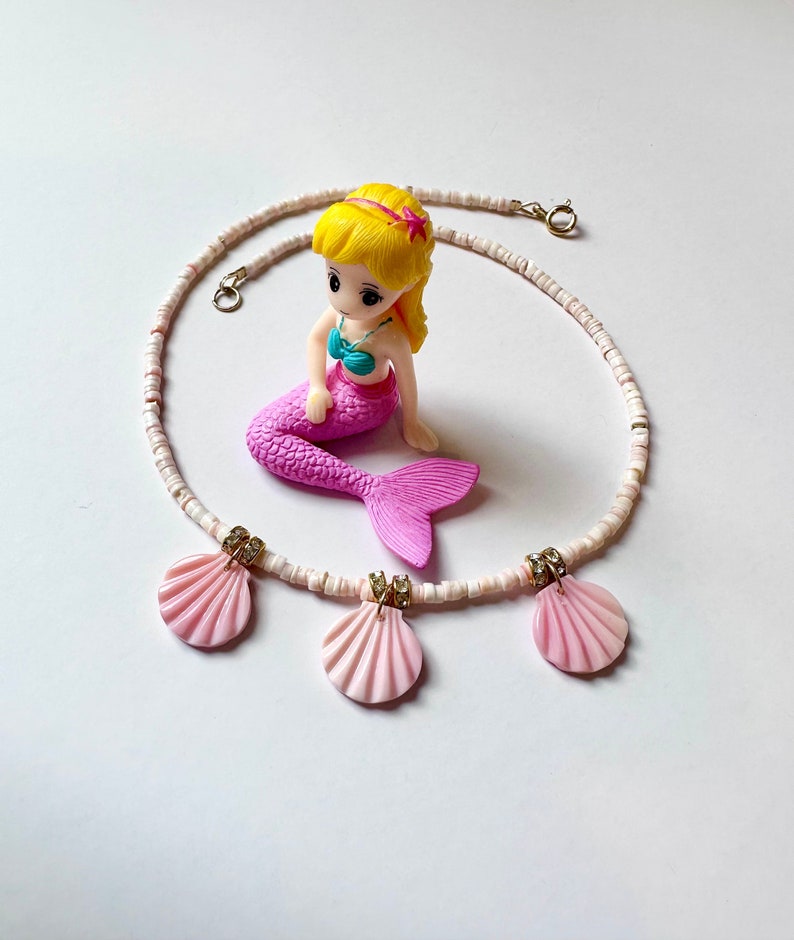 Puede incluir: Un collar de cuentas blancas con tres colgantes de conchas rosadas. El collar est&aacute; decorado con peque&ntilde;as cuentas doradas y diamantes de imitaci&oacute;n.