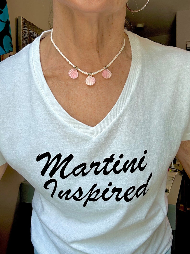 Puede incluir: Una camiseta blanca con una inscripci&oacute;n en cursiva negra que dice "Martini Inspired". La persona lleva un collar de cuentas blancas con tres colgantes de conchas rosas.