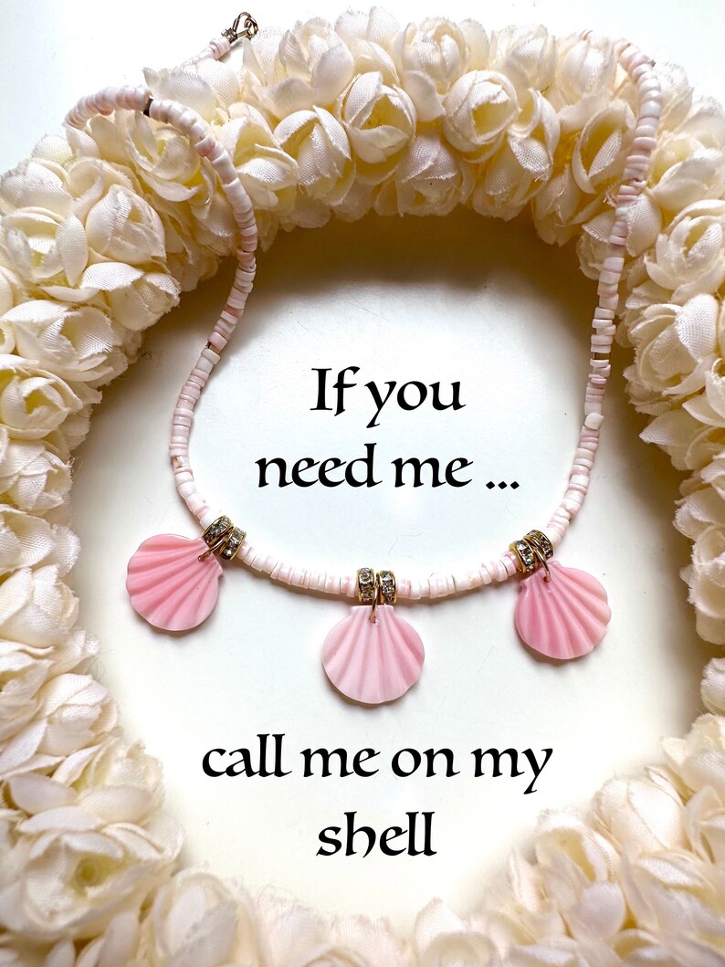 Puede incluir: Un collar de conchas blancas con colgantes de conchas rosas y diamantes de imitaci&oacute;n. El collar est&aacute; rodeado de una guirnalda de flores blancas. El texto "Si me necesitas... ll&aacute;mame a mi concha" est&aacute; escrito debajo del collar.