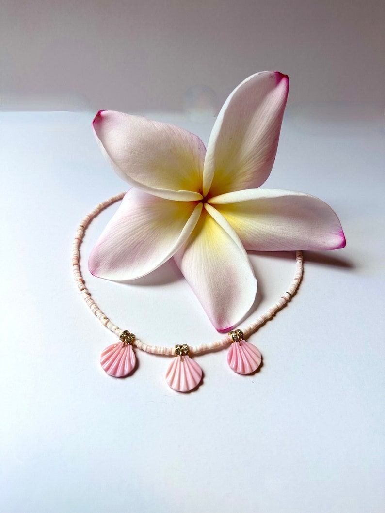 Puede incluir: Una delicada flor blanca y rosa con un collar de conchas rosa con tres colgantes de conchas rosas.