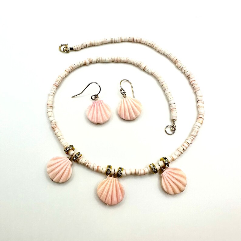 Puede incluir: Un collar de conchas blancas con tres colgantes de conchas rosas y un juego de pendientes a juego de conchas rosas. El collar tiene peque&ntilde;as cuentas blancas y diamantes de imitaci&oacute;n.