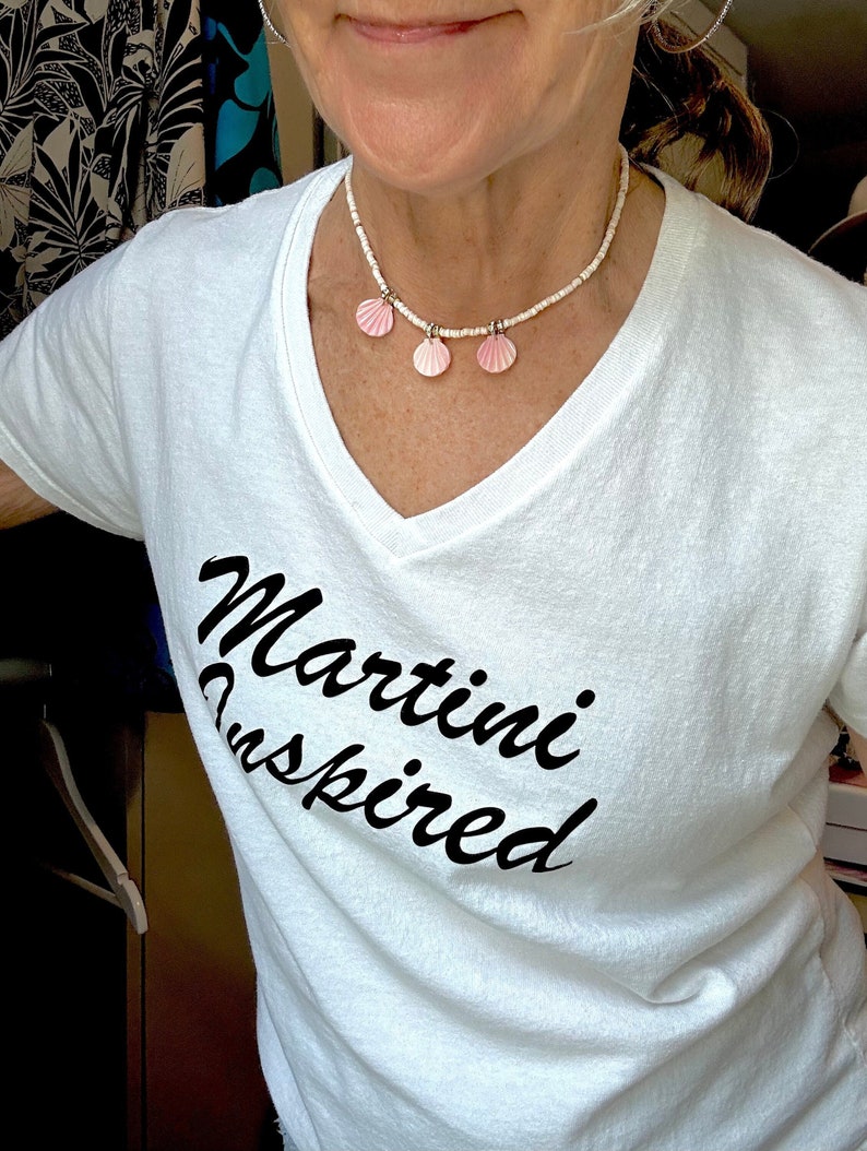 Puede incluir: Camiseta blanca con el texto "Martini Inspired" en escritura cursiva negra. La persona lleva un collar de cuentas blancas con tres dijes de conchas rosadas.