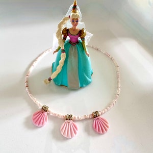 Puede incluir: Un collar de cuentas blancas con tres colgantes de concha rosa. El collar est&aacute; sobre una superficie blanca con una mu&ntilde;eca en el fondo.
