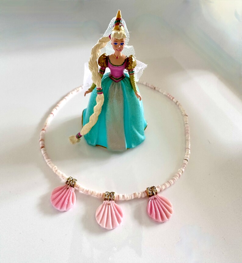 Puede incluir: Un collar de cuentas blancas con tres colgantes de concha rosa. El collar est&aacute; sobre una superficie blanca con una mu&ntilde;eca en el fondo.