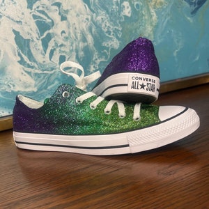 May include: Green and purple glitter Converse All Star trainers with white soles and black laces. The shoes are on a wooden surface.