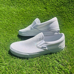May include: A pair of white slip-on sneakers with a glittery texture. The shoes are displayed on a bed of green grass, showcasing their clean design and casual style. The sneakers have a white sole and a simple, classic silhouette.