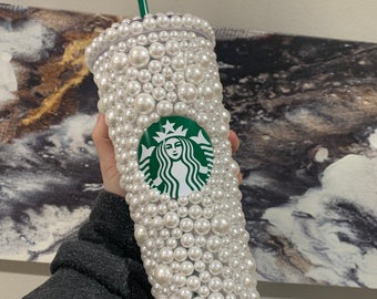 May include: A Starbucks cold cup, embellished with white faux pearls. The cup features the Starbucks logo in green and white. The lid and straw are green. The cup is held against a neutral background.