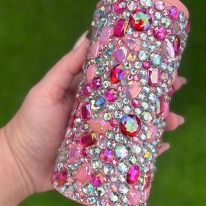 May include: A pink can cooler bedazzled with a variety of colorful rhinestones. The rhinestones are in shades of pink, red, and iridescent colors. The top of the cooler is silver and the rim is pink.