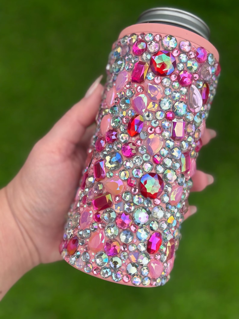 May include: A pink can cooler bedazzled with a variety of colorful rhinestones. The rhinestones are in shades of pink, red, and iridescent colors. The top of the cooler is silver and the rim is pink.