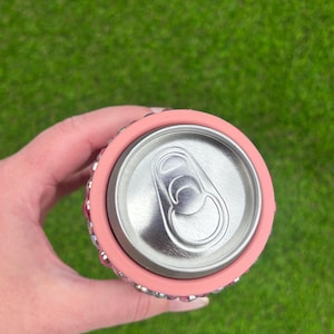 May include: A pink can cooler with a silver can inside. The cooler is decorated with small, sparkling rhinestones around the top and bottom edges. The background is a blurred green lawn.