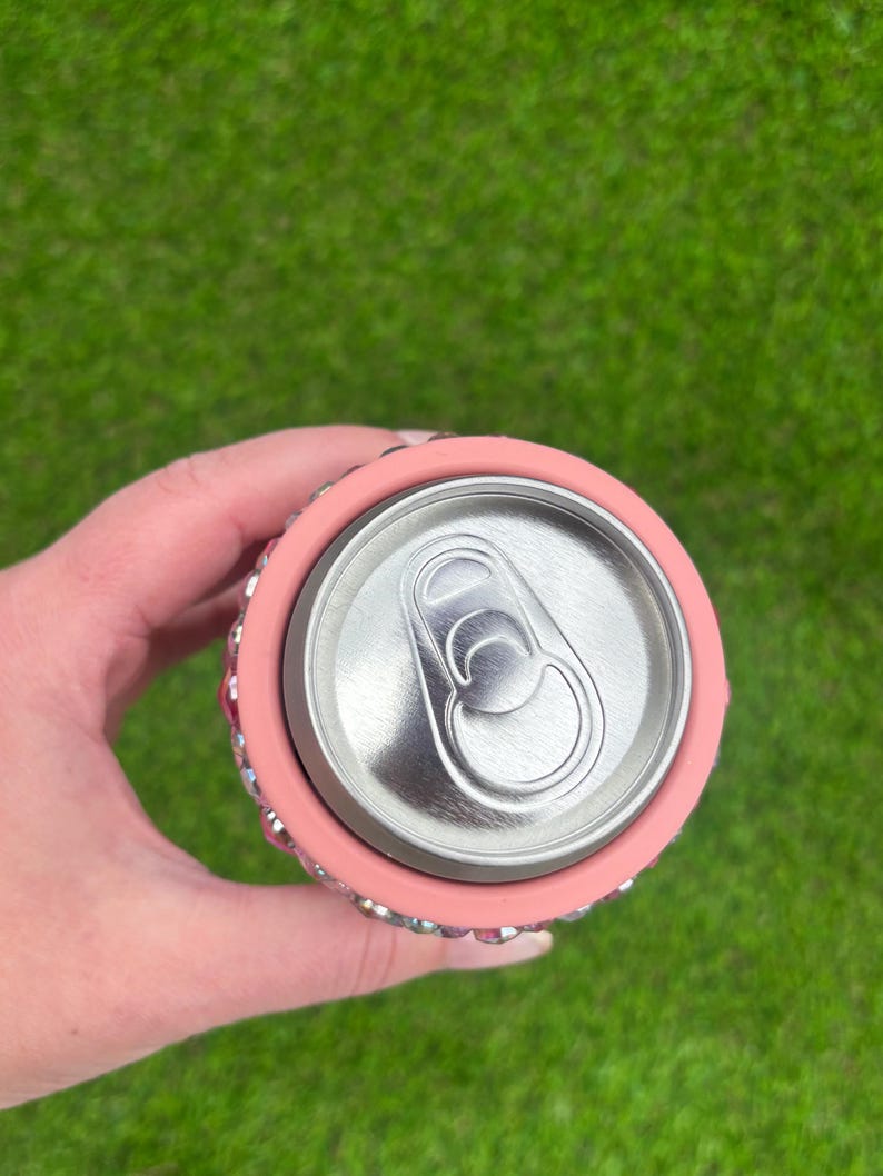 May include: A pink can cooler with a silver can inside. The cooler is decorated with small, sparkling rhinestones around the top and bottom edges. The background is a blurred green lawn.