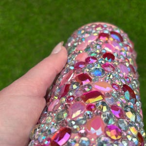 May include: A pink tumbler covered in a variety of iridescent, pink, and red rhinestones. The rhinestones are different shapes and sizes, creating a sparkling effect. The tumbler is held by a hand.