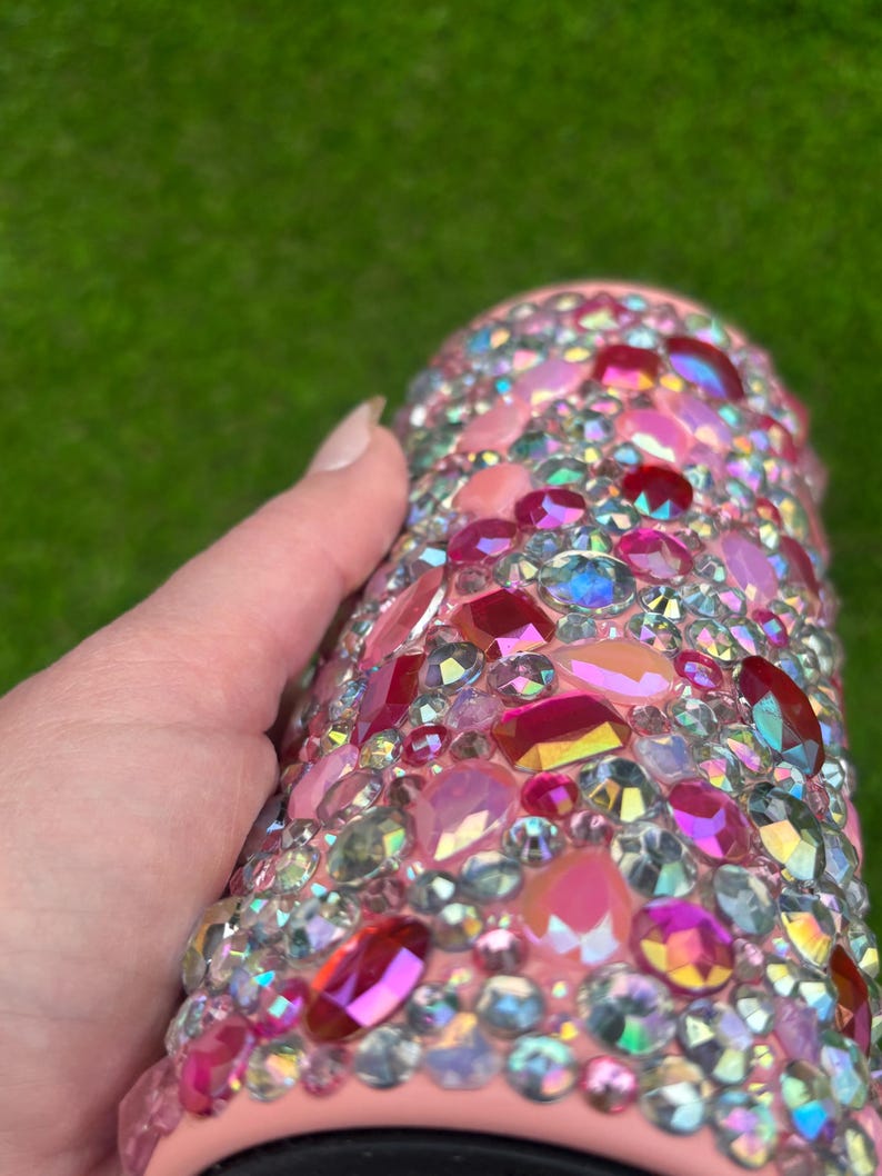 May include: A pink tumbler covered in a variety of iridescent, pink, and red rhinestones. The rhinestones are different shapes and sizes, creating a sparkling effect. The tumbler is held by a hand.