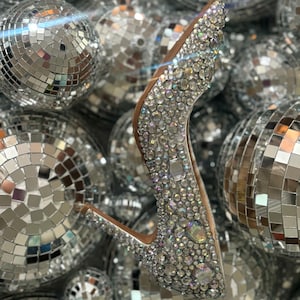 Op de afbeelding: Een zilveren hoge hak schoen bedekt met iriserende strass steentjes, omringd door zilveren discoballen.