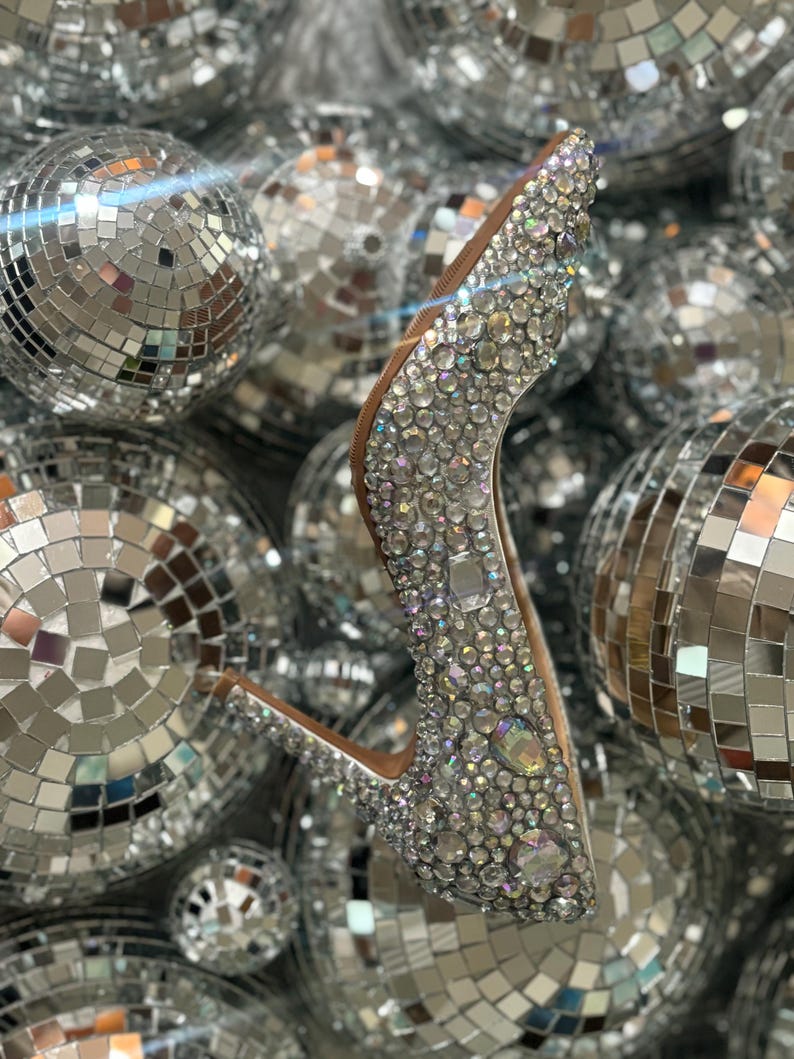 Op de afbeelding: Een zilveren hoge hak schoen bedekt met iriserende strass steentjes, omringd door zilveren discoballen.