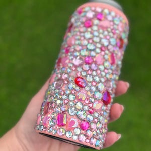May include: A pink insulated tumbler covered in a variety of sparkling rhinestones in shades of pink, red, and iridescent colors. The tumbler has a black base and a pink rim. The rhinestones are of different shapes and sizes, creating a dazzling effect.