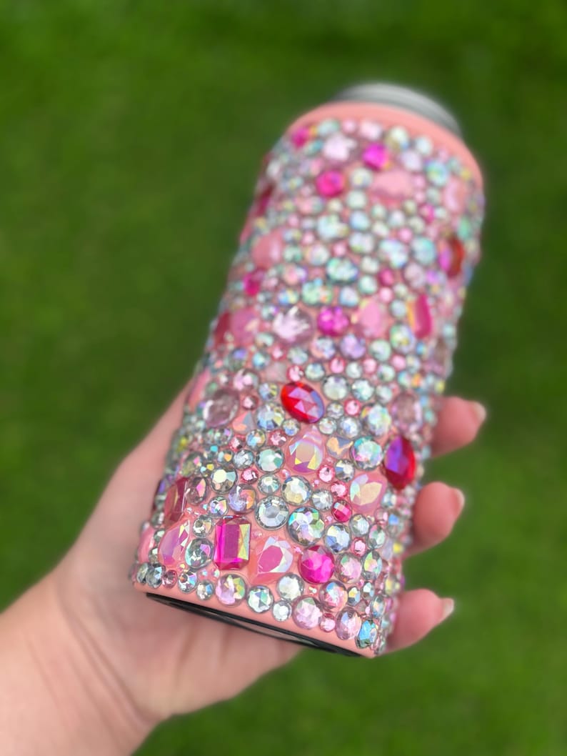 May include: A pink insulated tumbler covered in a variety of sparkling rhinestones in shades of pink, red, and iridescent colors. The tumbler has a black base and a pink rim. The rhinestones are of different shapes and sizes, creating a dazzling effect.