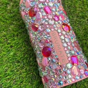 May include: A pink insulated can cooler bedazzled with a variety of pink, red, and clear rhinestones. The word "BR&Uuml;MATE" is printed vertically on the side of the cooler.