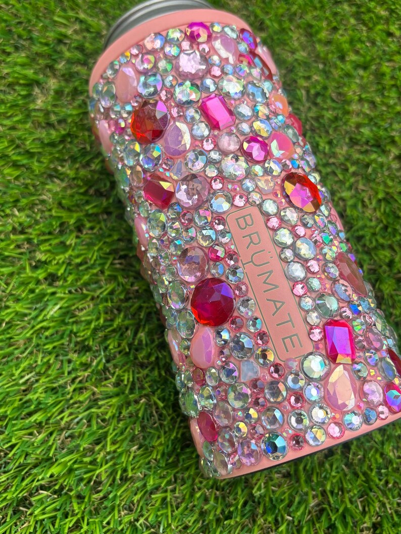May include: A pink insulated can cooler bedazzled with a variety of pink, red, and clear rhinestones. The word "BR&Uuml;MATE" is printed vertically on the side of the cooler.