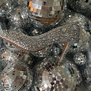 Op de afbeelding: Een zilveren hoge hak schoen bedekt met glinsterende strass steentjes, omringd door discoballen.