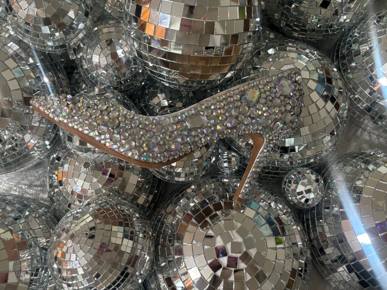 Op de afbeelding: Een zilveren hoge hak schoen bedekt met glinsterende strass steentjes, omringd door discoballen.