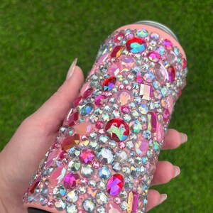 May include: A pink tumbler covered in a variety of iridescent, pink, and red rhinestones. The bedazzled tumbler has a light pink base and a black bottom. The background is a blurred green.
