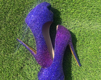 Op de afbeelding: Een paar pumps met hoge hakken en open teen, bedekt met een levendige paarse glitterafwerking. De schoenen zijn voorzien van een plateauzool en een dunne stilettohak, met een kleurverloop dat overgaat van dieppaars bij de teen naar een lichtere tint.