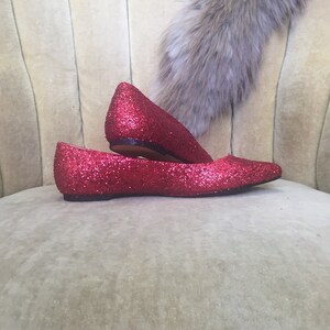 May include: A pair of red glitter flats with pointed toes. The shoes are sitting on a light-colored fabric surface.
