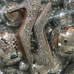Op de afbeelding: Een paar zilveren pumps met hoge hakken, bedekt met glinsterende strasssteentjes. De schoenen zijn gefotografeerd tegen een achtergrond van zilveren discoballen.