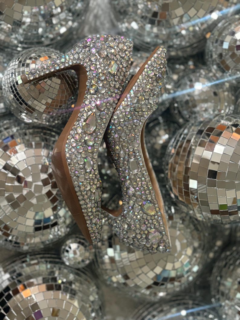 Op de afbeelding: Een paar zilveren pumps met hoge hakken, bedekt met glinsterende strasssteentjes. De schoenen zijn gefotografeerd tegen een achtergrond van zilveren discoballen.
